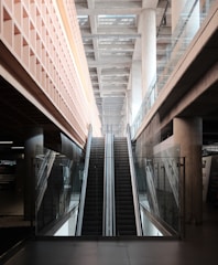 An indoor escalator ascends between two levels, with modern architectural features including glass railings and concrete pillars. The ceiling above is made of beams, providing a sense of depth and openness.