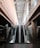An indoor escalator ascends between two levels, with modern architectural features including glass railings and concrete pillars. The ceiling above is made of beams, providing a sense of depth and openness.