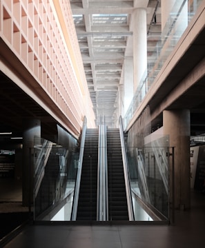 An indoor escalator ascends between two levels, with modern architectural features including glass railings and concrete pillars. The ceiling above is made of beams, providing a sense of depth and openness.