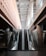 An indoor escalator ascends between two levels, with modern architectural features including glass railings and concrete pillars. The ceiling above is made of beams, providing a sense of depth and openness.