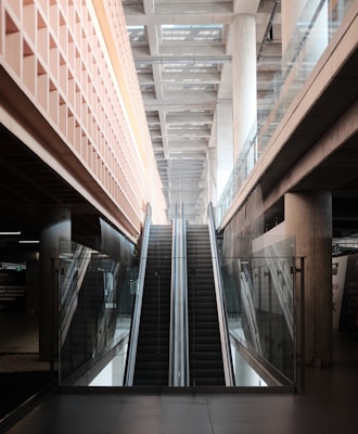 An indoor escalator ascends between two levels, with modern architectural features including glass railings and concrete pillars. The ceiling above is made of beams, providing a sense of depth and openness.