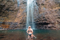 A person in a black swimsuit stands in a body of water at the base of a tall waterfall. The surrounding environment is rocky, with wet stones and moss. The water flows down from a significant height, creating a serene and natural atmosphere.