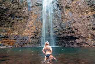 A person in a black swimsuit stands in a body of water at the base of a tall waterfall. The surrounding environment is rocky, with wet stones and moss. The water flows down from a significant height, creating a serene and natural atmosphere.