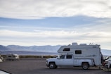 A slide-in camper mounted on a truck, set against a backdrop of dense Alaskan forest and clear blue sky.