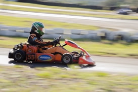 A person in a green helmet and orange racing suit is driving a go-kart on a racetrack. The go-kart is predominantly orange with the number 55 and the brand name 'Skinabon' visible on the side. The background is blurred, indicating speed, and the track is surrounded by grass and white barriers.