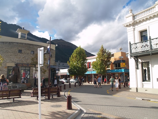 A bustling street scene with various shops and buildings lining the road. People are walking around, enjoying a sunny day. The street is paved, with benches and trees providing a cozy urban atmosphere. A mountain looms in the background under a partly cloudy sky.