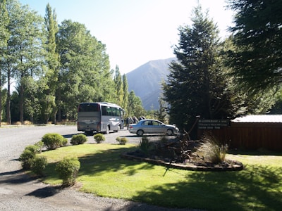 A scenic area with a lush green lawn bordered by shrubs and trees. A large tour bus and a car are parked on a gravel driveway. Tall trees provide a backdrop with mountains visible in the distance. There is a wooden sign indicating the presence of a restaurant or lodge nearby.