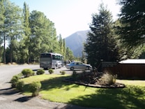 A scenic area with a lush green lawn bordered by shrubs and trees. A large tour bus and a car are parked on a gravel driveway. Tall trees provide a backdrop with mountains visible in the distance. There is a wooden sign indicating the presence of a restaurant or lodge nearby.