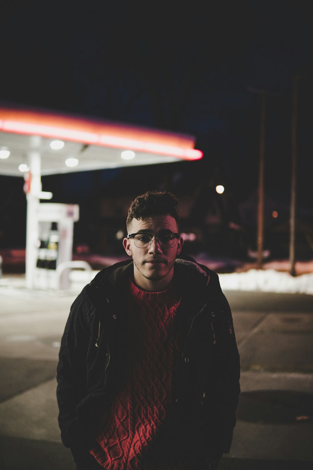 man in red shirt and black zip-up hooded jacket standing near white and red gas station at night