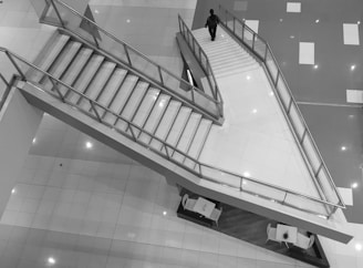 An overhead view of a modern staircase in a building, featuring clean lines and sleek metal railings. A single person is descending the stairs. Below the stairs, a seating area with tables and chairs is visible. The floor is tiled with a glossy finish, creating a reflective surface.