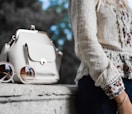 woman wearing beige and red floral top leaning on gray concrete slab with white leather bag ontop