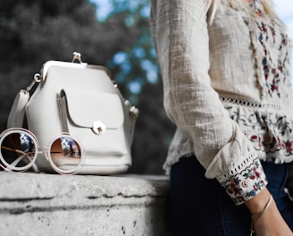 woman wearing beige and red floral top leaning on gray concrete slab with white leather bag ontop