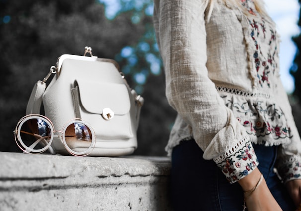 woman wearing beige and red floral top leaning on gray concrete slab with white leather bag ontop