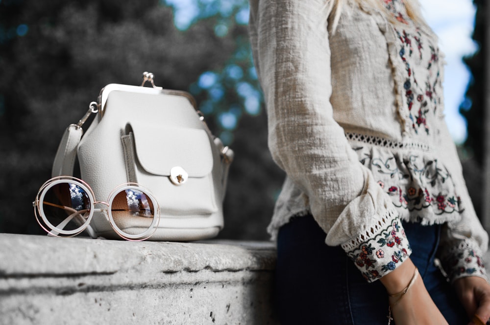 woman wearing beige and red floral top leaning on gray concrete slab with white leather bag ontop