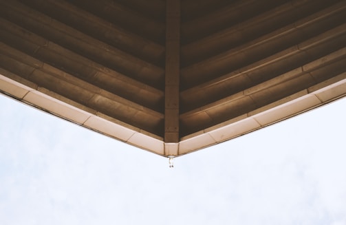 The image captures the underside of a large roof structure with a pointed design. The roof appears to be made of concrete or a similar material, with visible beams and lines running parallel to the point. The sky is visible in the background, offering a contrast to the architectural element.