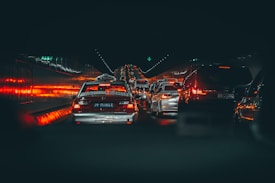 A congested traffic scene in a tunnel with numerous vehicles, including taxis and cars, lined up and emitting red brake lights that illuminate the sides of the tunnel. The atmosphere is dimly lit, with a focus on the glowing lights reflecting off the wet road and tunnel walls.