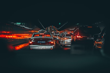 A congested traffic scene in a tunnel with numerous vehicles, including taxis and cars, lined up and emitting red brake lights that illuminate the sides of the tunnel. The atmosphere is dimly lit, with a focus on the glowing lights reflecting off the wet road and tunnel walls.