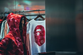 A person wearing a red camo hoodie stands near a clothing rack with several hangers displaying t-shirts. One of the shirts has a bold red graphic of a face. The lighting is dim and the setting appears to be a store or clothing boutique.
