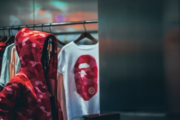 A person wearing a red camo hoodie stands near a clothing rack with several hangers displaying t-shirts. One of the shirts has a bold red graphic of a face. The lighting is dim and the setting appears to be a store or clothing boutique.