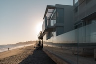A modern beachfront house with large glass windows reflecting a serene beach scene. The sun is setting on the horizon, casting a warm glow over the water and sand. A few people are walking along the shoreline, enjoying the peaceful atmosphere.