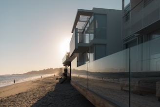 A modern beachfront house with large glass windows reflecting a serene beach scene. The sun is setting on the horizon, casting a warm glow over the water and sand. A few people are walking along the shoreline, enjoying the peaceful atmosphere.