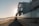 A modern beachfront house with large glass windows reflecting a serene beach scene. The sun is setting on the horizon, casting a warm glow over the water and sand. A few people are walking along the shoreline, enjoying the peaceful atmosphere.