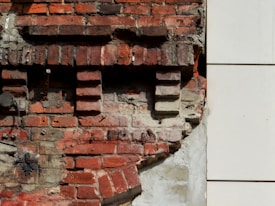 An old brick wall with visible signs of wear and tear, showing chipped bricks and patches of worn concrete. The bricks are arranged in a classic staggered pattern with areas of exposed mortar. To the right, there is a white, smooth surface contrasting with the rough texture of the bricks.