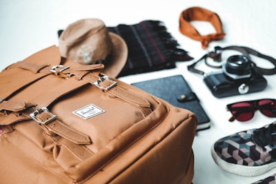 Various travel essentials are organized on a flat surface, including a tan backpack, a brown hat, a black notebook, a plaid scarf, a bandana, a vintage camera, sunglasses, and a patterned sneaker.