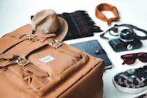 Various travel essentials are organized on a flat surface, including a tan backpack, a brown hat, a black notebook, a plaid scarf, a bandana, a vintage camera, sunglasses, and a patterned sneaker.