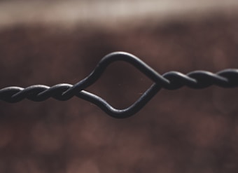 Close-up view of a twisted and looped metal wire against a blurred brown background. The wire's intricate twist forms a distinctive loop in the center.
