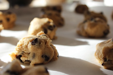 Close-up of raw chocolate chip cookie dough balls placed on a baking sheet, ready for baking. The dough is light brown with visible dark chocolate chunks, set in a sunlit environment creating soft shadows.