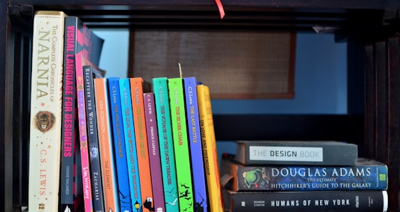 A bookshelf filled with various books, including a complete set of 'The Chronicles of Narnia' by C.S. Lewis, and titles like 'Visual Language for Designers' and 'The Hitchhiker's Guide to the Galaxy' by Douglas Adams. The books are neatly arranged, some placed vertically and others horizontally. The background features a wooden structure, adding a cozy and intellectual atmosphere.