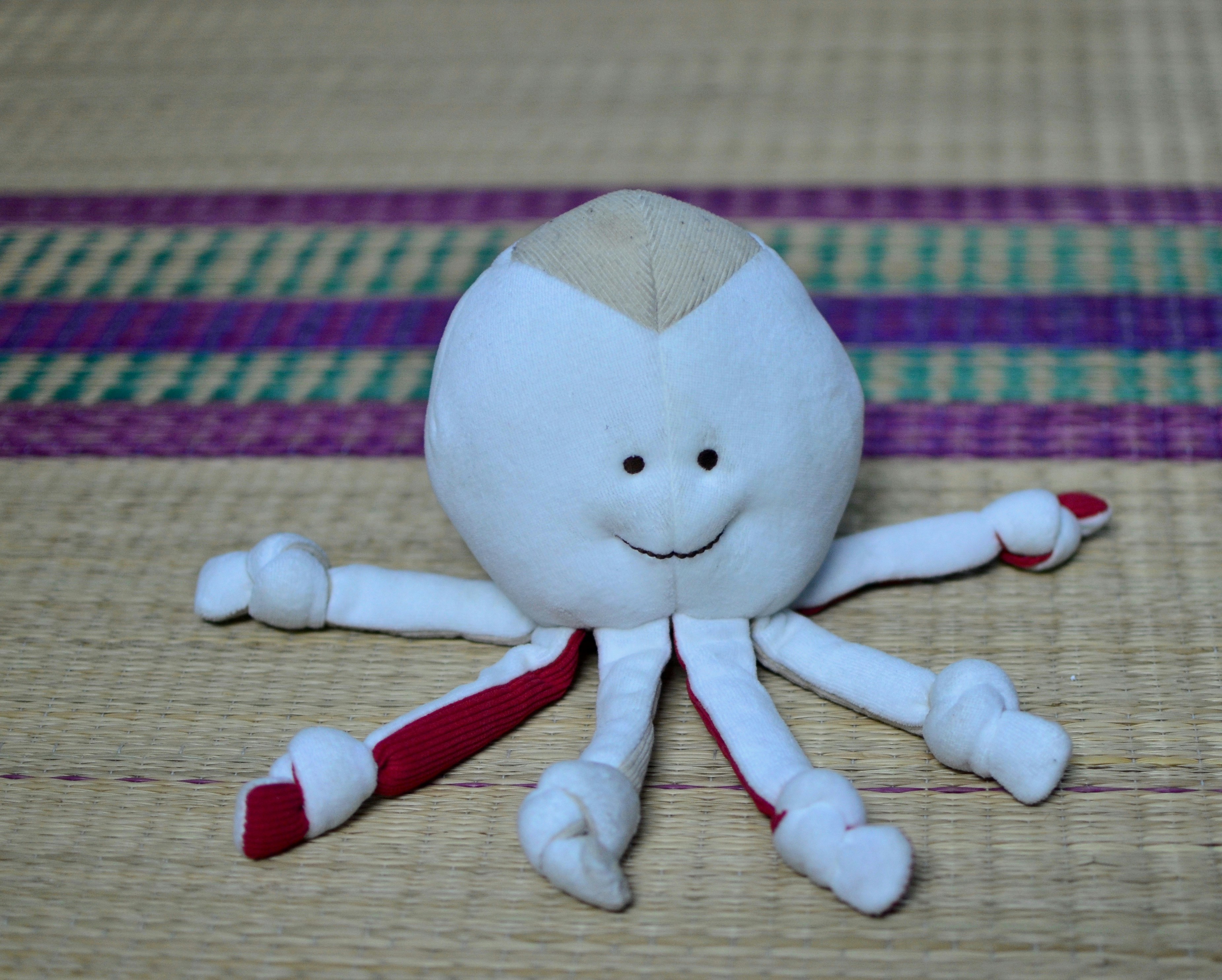 A plush toy resembling an octopus with a smiling face is positioned on a woven mat. The toy is predominantly white with red accents on its tentacles, which have small knots tied at the ends. The background features a woven mat with colorful patterns in purple and green.