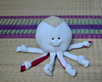 A plush toy resembling an octopus with a smiling face is positioned on a woven mat. The toy is predominantly white with red accents on its tentacles, which have small knots tied at the ends. The background features a woven mat with colorful patterns in purple and green.