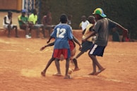 A group of children are playing soccer on a dirt field, surrounded by other children watching from the sidelines. The three main players are engaged in action, with one sporting a blue jersey marked with the number 12. The atmosphere is casual, with most players and spectators wearing informal clothing.