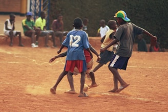 A group of children are playing soccer on a dirt field, surrounded by other children watching from the sidelines. The three main players are engaged in action, with one sporting a blue jersey marked with the number 12. The atmosphere is casual, with most players and spectators wearing informal clothing.