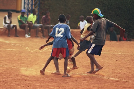 A group of children are playing soccer on a dirt field, surrounded by other children watching from the sidelines. The three main players are engaged in action, with one sporting a blue jersey marked with the number 12. The atmosphere is casual, with most players and spectators wearing informal clothing.