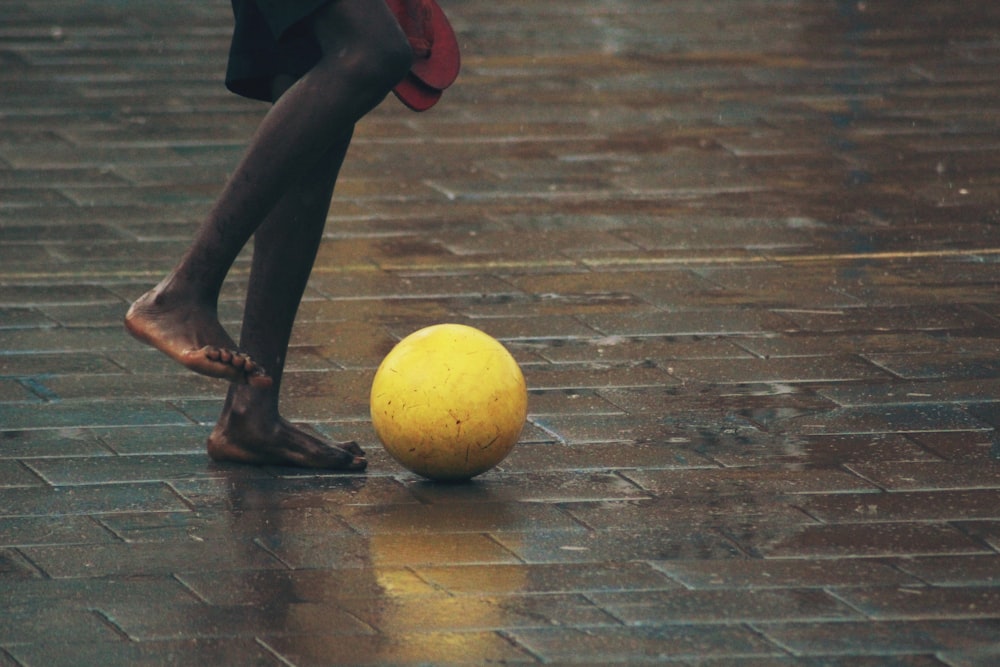 pessoa em pé chão de tijolo cinza prestes a chutar bola