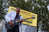 A musician is energetically playing an electric guitar while wearing a white jacket and sunglasses. He is outdoors in front of a sign that reads 'Independents' Day'. Trees are visible in the background, and the musician appears to be enjoying the performance.