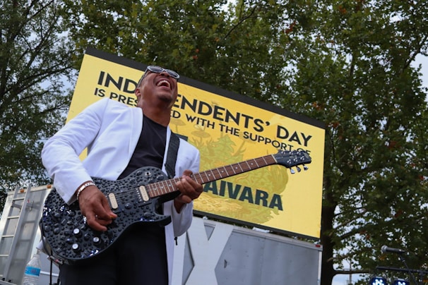 A musician is energetically playing an electric guitar while wearing a white jacket and sunglasses. He is outdoors in front of a sign that reads 'Independents' Day'. Trees are visible in the background, and the musician appears to be enjoying the performance.