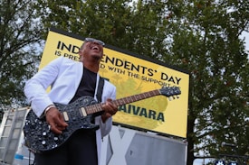 A musician is energetically playing an electric guitar while wearing a white jacket and sunglasses. He is outdoors in front of a sign that reads 'Independents' Day'. Trees are visible in the background, and the musician appears to be enjoying the performance.