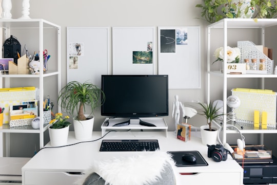 A tidy and modern workspace featuring a computer monitor, keyboard, and other office accessories. White shelving units house an array of books, plants, and decorative items, providing a clean and organized aesthetic. A mannequin figure and a variety of stationery cups add creative touches, while plants introduce a natural element.