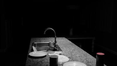 A dimly lit kitchen scene with a focus on a granite countertop featuring a modern faucet. Various kitchen items such as a stack of plates, a tin can, and a jar with a red lid are visible. The background is mostly dark, creating a contrast with the illuminated counter.