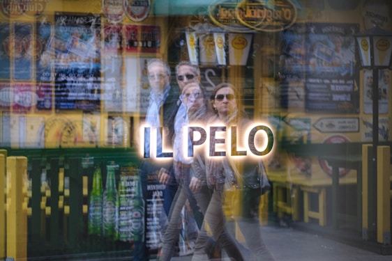 A reflection on glass showing an urban scene with people walking and various signs, advertisements, and posters in the background. The image features overlapping reflections, creating a dynamic and layered effect.