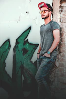A young man wearing a stylish cap and sunglasses, standing against a graffiti wall.