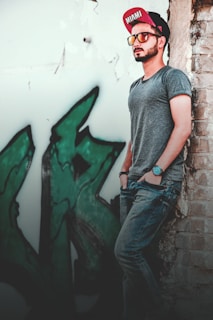 A stylish man adjusting his cap while leaning against a city wall