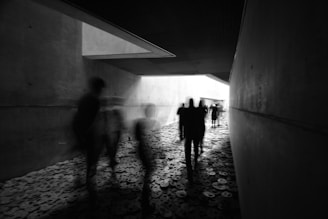 A dimly lit hallway with blurred figures walking over a floor covered in metal disks. The walls and floor have a concrete texture. The scene conveys an eerie and somber atmosphere with shadows cast by soft light entering from an opening at the far end.