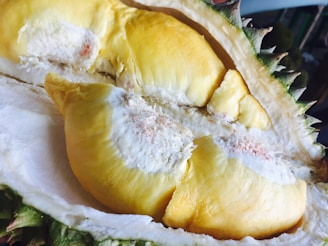 A close-up view of a cut-open durian fruit, revealing its creamy, yellow flesh surrounded by a spiky outer shell. The interior looks soft and smooth, contrasting with the rough texture of the rind.