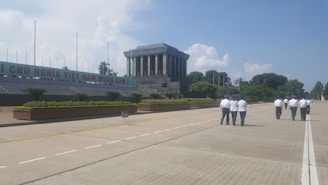 A large stone building with columns stands prominently in the center. A group of people dressed in uniforms are walking on a broad, empty boulevard. Lush green bushes and trees line the perimeter. Clear blue skies with fluffy clouds are visible in the background.
