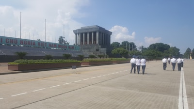 A large stone building with columns stands prominently in the center. A group of people dressed in uniforms are walking on a broad, empty boulevard. Lush green bushes and trees line the perimeter. Clear blue skies with fluffy clouds are visible in the background.